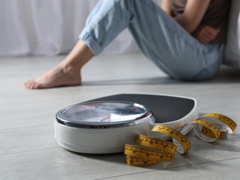 Eating disorder. Sad woman sitting near scale and measuring tape on floor indoors